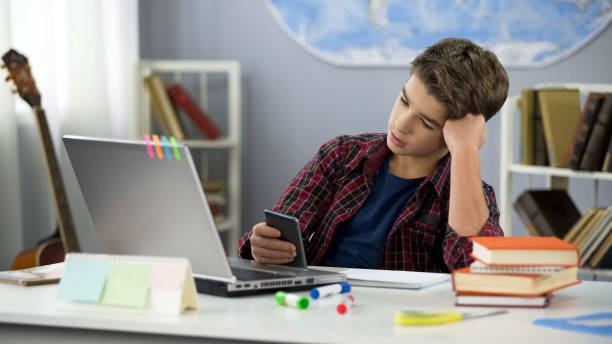 chico aburrido de desplazamiento aplicaciones en el teléfono, distraído de la tarea, procrastinación - perder el tiempo fotografías e imágenes de stock