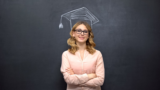 Smart girl stands against academic cap picture on blackboard, education program