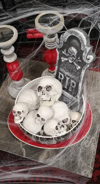 Photo of Human skulls stacked in dish with tombstone rest in peace with skull and bones behind,surrounded by empty candlesticks,on square gray tabletop,covered in cobwebs,with red-bound books in the background
