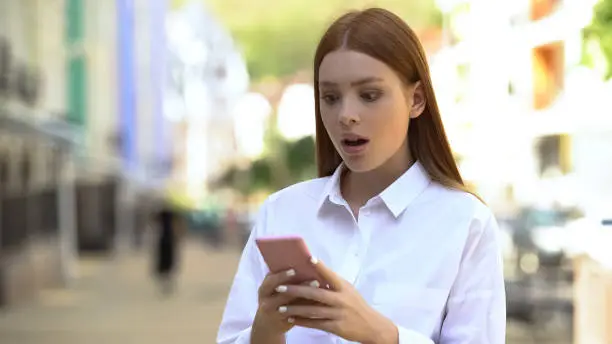 Young redhead female scrolling social network on smartphone, shocked by news