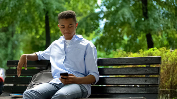 giovane adolescente che scorre smartphone, seduto in panchina nel parco, procrastina - women dependency business time foto e immagini stock