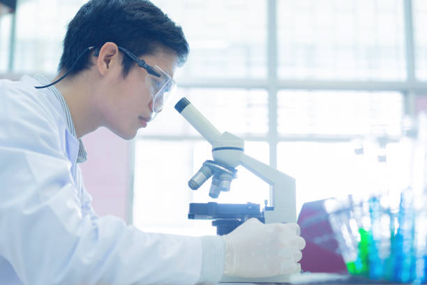 クローズアップアジアの科学者の男性は、医療と薬の概念に関する研究開発のための研究室で化学物質について分析するために顕微鏡を見て試みて制服を着用 - リサーチ ストックフォトと画像