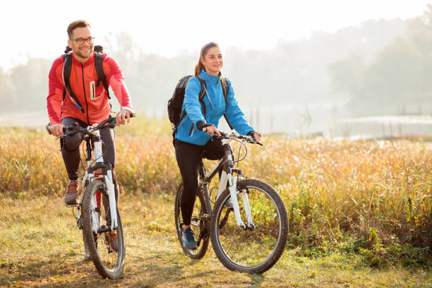 Beautiful happy young couple enjoying early morning bicycle ride by the river or lake Beautiful happy young couple enjoying early morning bicycle ride by the river or lake. Sunrise through the mist above water in the background red white blue bike stock pictures, royalty-free photos & images