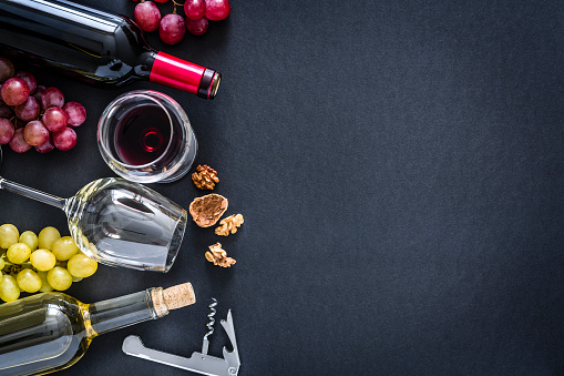 Top view of a black background with a composition of red, rosé and white wine bottles and wineglasses arranged at the left border leaving useful copy space for text and/or logo at the right. Red and white grapes and a vintage corkscrew complete the composition. Predominant colors are red, green and black. XXXL 42Mp studio photo taken with Sony A7rii and Sony FE 90mm f2.8 macro G OSS lens