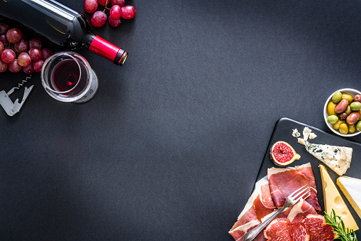 Appetizer: top view of a black background with a composition of red wine bottle, wineglass, a cutting board with various cheeses and Iberico ham arranged at the top left and bottom right corners leaving useful copy space for text and/or logo at the center. Some grapes, olives and a corkscrew complete the composition. Predominant colors are red and black. XXXL 42Mp studio photo taken with Sony A7rii and Sony FE 90mm f2.8 macro G OSS lens