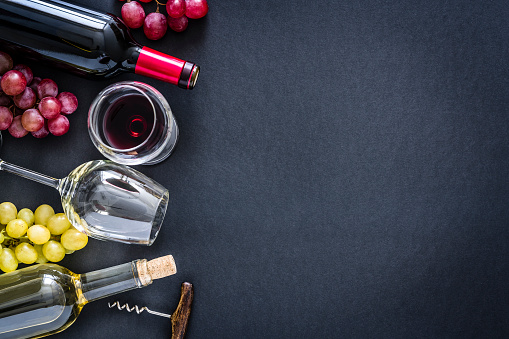 Top view of a black background with a composition of red, rosé and white wine bottles and wineglasses arranged at the left border leaving useful copy space for text and/or logo at the right. Red and white grapes and a vintage corkscrew complete the composition. Predominant colors are red, green and black. XXXL 42Mp studio photo taken with Sony A7rii and Sony FE 90mm f2.8 macro G OSS lens