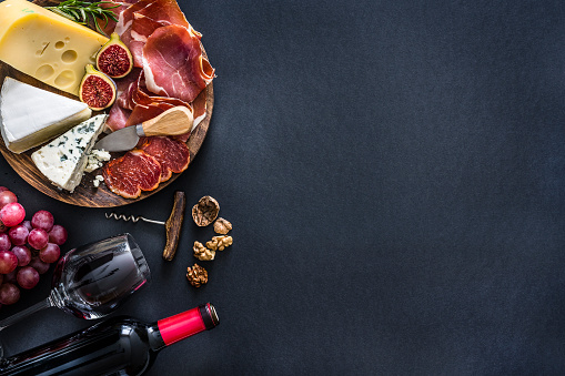 Appetizer: top view of a black background with a composition of red wine bottle, wineglass, a cutting board with various cheeses and Iberico ham arranged at the left border leaving useful copy space for text and/or logo at the right. Some grapes, nuts and a corkscrew complete the composition. Predominant colors are red and black. XXXL 42Mp studio photo taken with Sony A7rii and Sony FE 90mm f2.8 macro G OSS lens