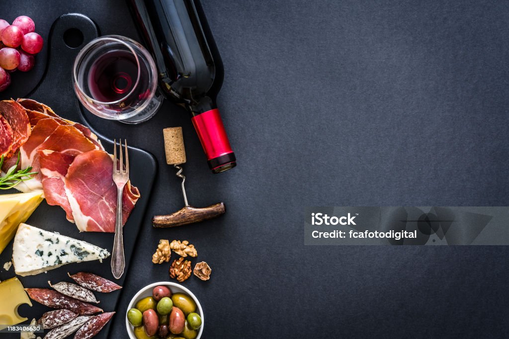Appetizer frame: red wine, Iberico ham and cheese on rustic table Appetizer: top view of a black background with a composition of red wine bottle, wineglass, a cutting board with various cheeses and Iberico ham arranged at the left border leaving useful copy space for text and/or logo at the right. Some grapes, olives, nuts and a corkscrew complete the composition. Predominant colors are red and black. XXXL 42Mp studio photo taken with Sony A7rii and Sony FE 90mm f2.8 macro G OSS lens Wine Stock Photo