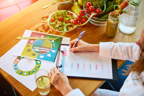 mujer dietista en uniforme médico con cinta métrica trabajando en un plan de dieta sentado con diferentes ingredientes alimenticios saludables en el consultorio verde sobre el fondo. pérdida de peso y concepto de nutrición adecuado - prescription plan fotografías e imágenes de stock