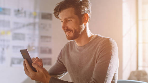 istock Professional Creative Man Sitting at His Desk in Home Office Studio Using Smartphone, Man Using Mobile Phone, Browsing Through Internet, Social Networks / Media. 1183411325