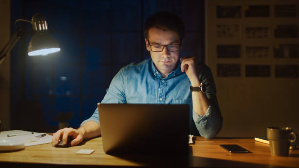 istock Professional Man Sitting at His Desk in Office Studio Working on a Laptop in the Evening. Man Analyzing Statistics. 1183411351