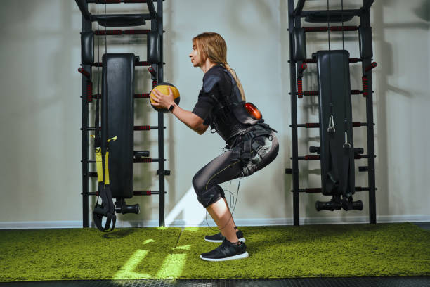 Beautiful blonde woman wearing in sensors and belts, fastens with velcro is practicing EMS fitness in a gym, building up muscles with a weighted ball. Modern kind of sport stock photo