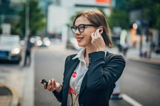 Businesswoman using wireless in ear headphones and mobile phone while walking on the street
