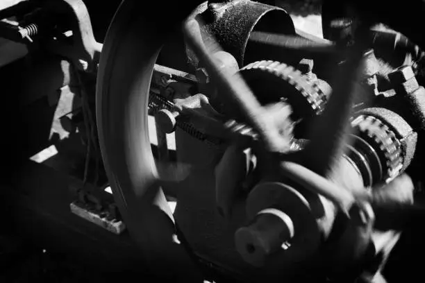 A vintage flywheel water pump engine appears close-up, in black and white, in a horizontal composition showing the main wheel, gearing and piston apparatus.