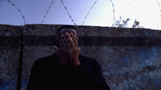 Depressed afro-american man standing near barbed wire fence, regretting crime