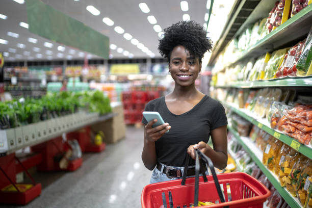 porträtt av leende kvinna med hjälp av telefon och hålla en varukorg i supermarket - kundkorg bildbanksfoton och bilder