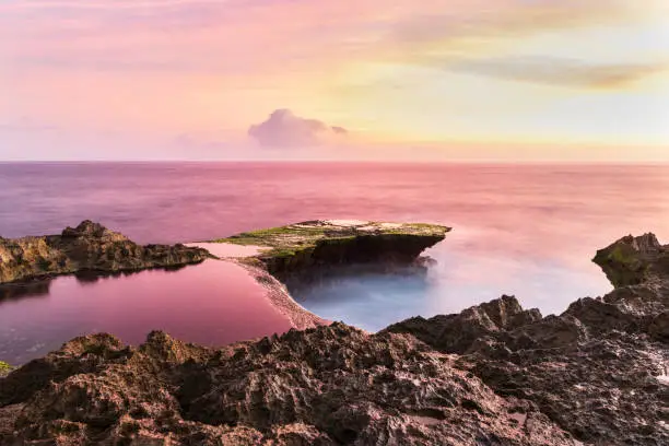Devil's Tear at sunset, island of Nusa Lembongan, Bali, Indonesia. Rocky shore in foreground. Tidepool and ocean pink from setting sun's reflection. Pink and yellow sky beyond.