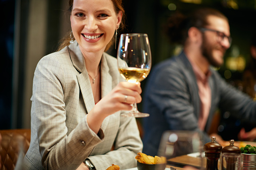 Portrait of beautiful caucasian brunette dressed elegant posing with glass of wine while sitting in restaurant with her friends.
