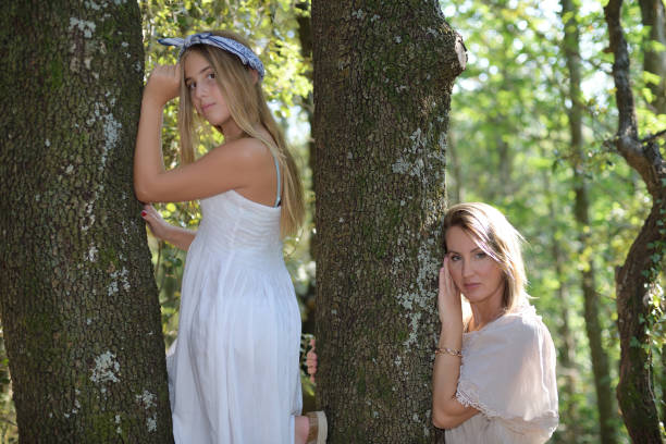 Mother and daughter leaning on a tree in a forest with clear dresses and looking at the camera