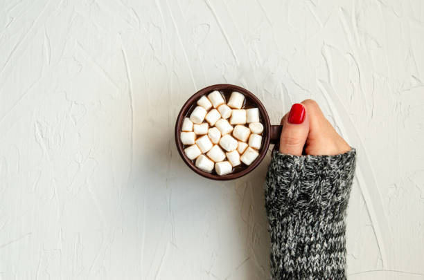 istock Female hand holds a brown cup with hot cocoa with white marshmallows on a white background with space for copy. 1183456971