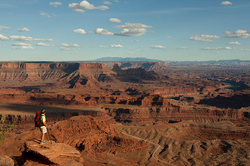 Young man wearing backpack standing on huge rocky ledge looking at view of Canyonlands National Park in Moab, Utah with blue sky in the foreground.
