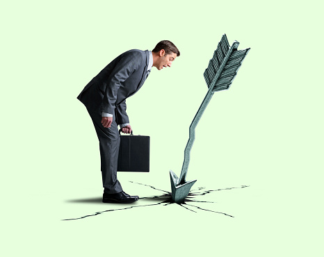 A businessman stands while holding his briefcase as he looks down at a large arrow and the hole and cracks in the ground it has created isolated against a light green background.