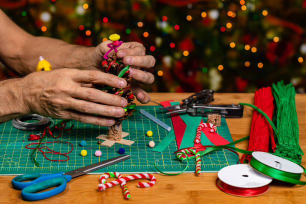 Someone Making holiday crafts. A Christmas Tree being created from a large Pine cone with ribbon, bells and pompoms. There is negative space for Copywriting on the soft focused background full of heavy bokeh holiday lights alguien haciendo manualidades navideñas. un árbol de navidad que se crea a partir de un gran cono de pino con cinta, campanas y pompones. hay espacio negativo para la escritura de copias en el fondo de enfoque suave lleno de luces de vacaciones bokeh p
