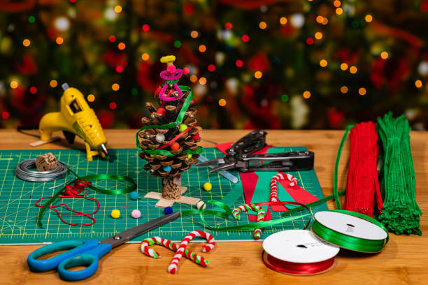 istock Making holiday crafts.  A Christmas Tree being created from a large Pine cone with ribbon, bells and pompoms.  There is negative space for Copywriting on the soft focused background full of heavy bokeh holiday lights 1183511184