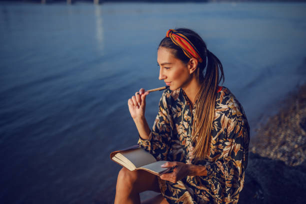 porträt der nachdenklichen attraktiven kaukasischen brünette in floralen kleid und mit stirnband sitzen auf felsen am ufer neben dem fluss und tagebuch schreiben. - nature writing women ideas stock-fotos und bilder