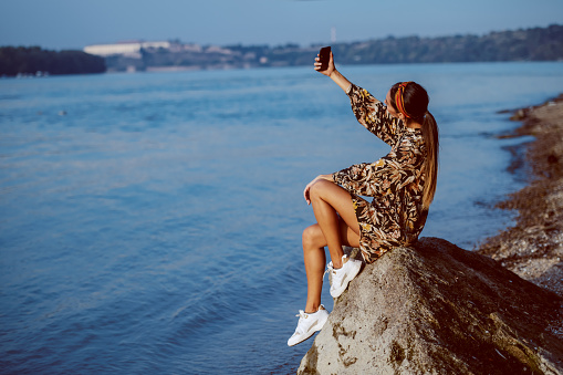 Good-looking caucasian brunette in floral dress and with headband sitting on the rock on shore next to river and taking selfie.
