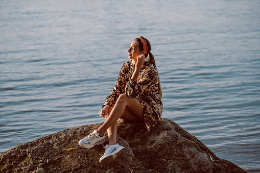 Full length of beautiful caucasian brunette with headband and in floral dress sitting on rock next to river, enjoying sunny day and listening music.