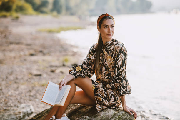 encantadora morena caucásica con diadema y vestido floral sentado en roca cerca del río, sosteniendo el libro y mirando hacia otro lado. - floral dress fotografías e imágenes de stock