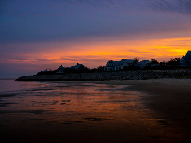 The last rays of brilliant orange and pink sun smudge the sky and are reflected in the calm water washing up on the beach along the coast of Cape Cod