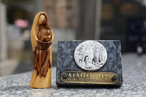 Photo of France. April, 26, 2019. Statue of the Virgin and Child in olive wood placed on a tombstone and close to a commemorative plaque composed of an inscription: to the cave bless, I prayed for you.