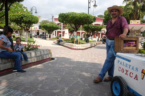 Aquismon, Mexico, September 08, 2018: Icecream vendor with a cowboy hat and moustache in the square with buildings in the background and a mother and child sitting across the street. Travel concept