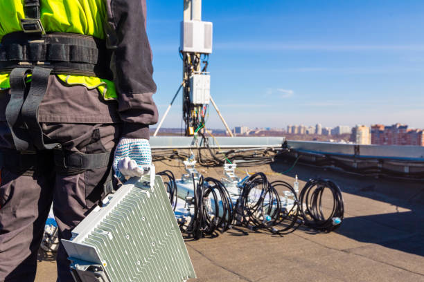 Professional industrial climber in uniform with telecomunication equipment in his hand and antennas of GSM DCS UMTS LTE bands, outdoor radio units on the roof. Working process of upgrading or swap telecommunication equipment escalador industrial profesional en uniforme con equipo de telecomunicaciones en su mano y antenas de bandas gsm dcs umts lte, unidades de radio al aire libre en el techo. proceso de trabajo de actualización o intercambio de equipos de telecomunicaciones.