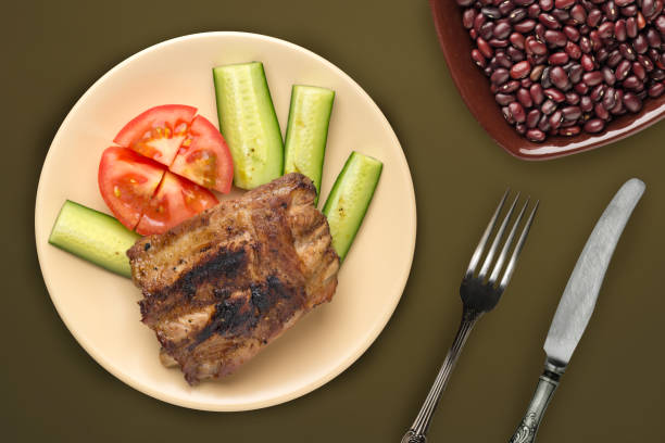 fried pork ribs with chopped cucumbers and tomatoes on a beige plate. pork ribs on an brown green background. grilled ribs