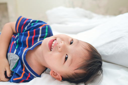 Cute little happy smiling Asian 3 - 4 years toddler boy child waking up in bed, Cheerful kid lying on bed looking at camera on lazy day, Morning awakening, Good morning at home concept