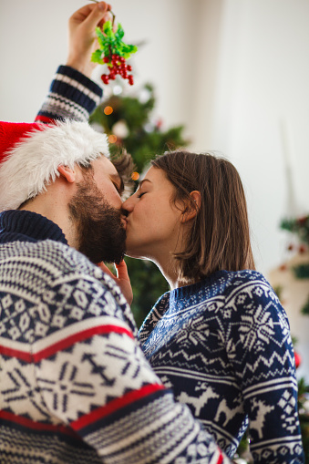 Affectionate young couple wearing cozy Christmas jumpers standing under mistletoe and kissing on Christmas day.