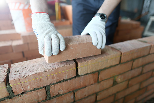 Close-up of prudent man wearing special protective gloves to prevent serious work injuries. Male hands putting brick on wall on big masonry. Building concept