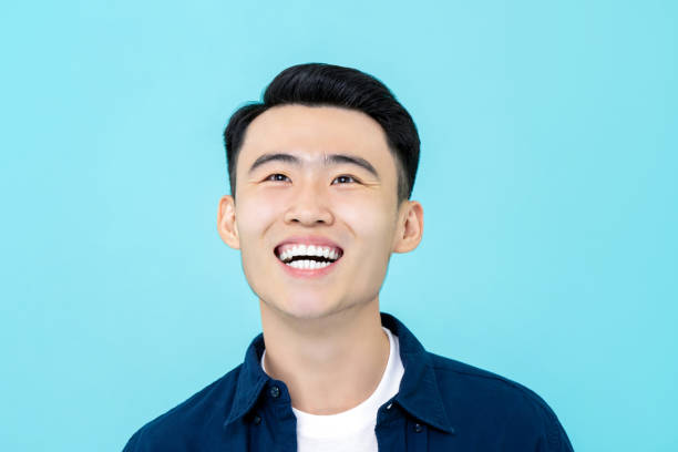 Handsome happy young Asian man smiling and looking upwards Happy young Asian man smiling and looking upwards isolated on light blue background admired stock pictures, royalty-free photos & images