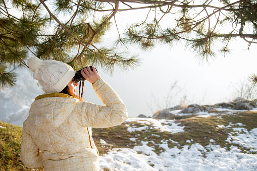 The girl in a White cap with a backpack in the winter in a forest looking through binoculars