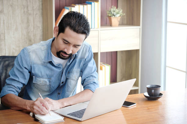 istock Owner Business or Entrepreneur using a laptop for working and writing work schedules in the notebook while sitting at the desk in his home. 1183699081