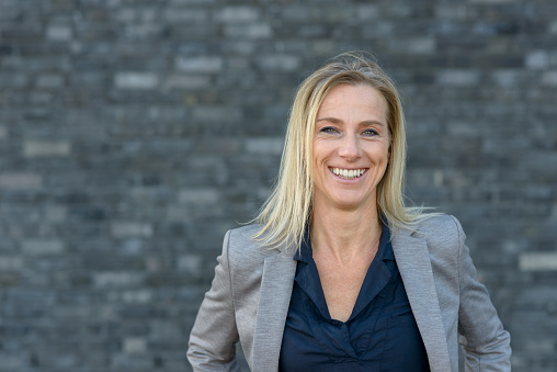 Front portrait of business woman with blond hair, standing outdoors, looking at camera and smiling. Grey background copy space