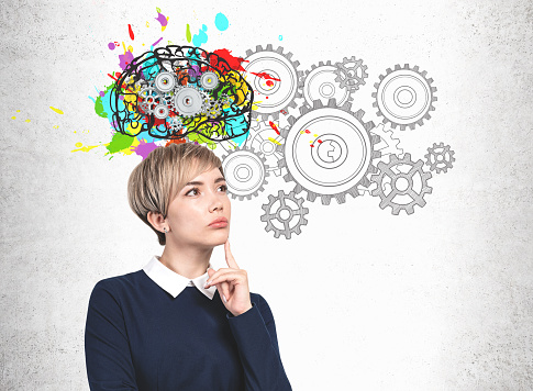 Pensive blonde young woman in blue dress standing near concrete wall with colorful brain sketch with gears drawn on it. Concept of brainstorming and creative thinking