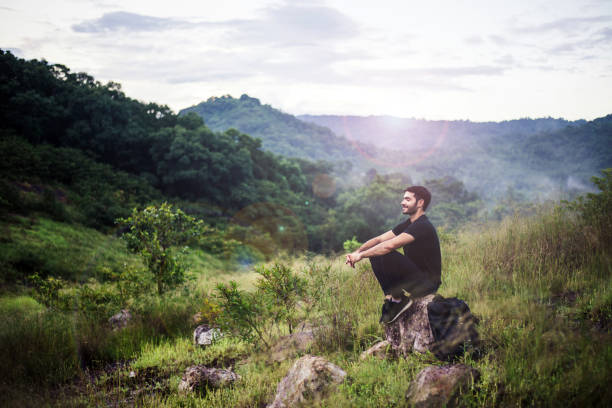 viaggiatore con zaino seduto sulla roccia all'aperto, meditazioni per calmare la mente e il corpo - sunset yoga young men caucasian foto e immagini stock