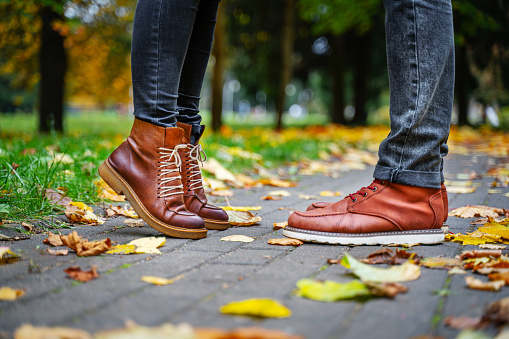 Feet of a couple in love in brown shoes on the path of the autumn park, strewn with fallen leaves. Girl stands on toes. Kiss concept