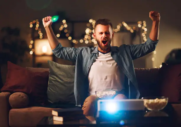 young male cheerleader watching football on tv projector at home in the evening alone