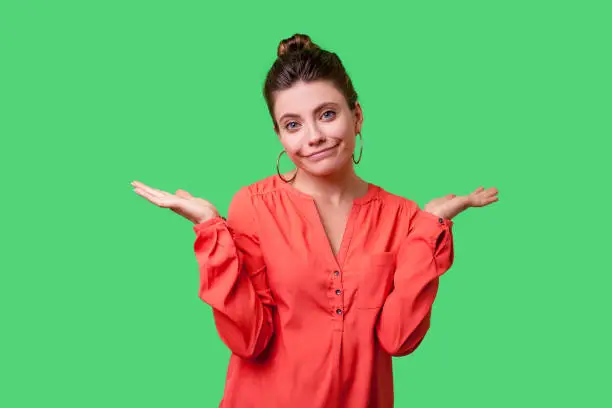 Not sure! Portrait of uncertain confused woman with bun hairstyle, big earrings and in red blouse shrugging shoulders, showing i don't know gesture. indoor studio shot isolated on green background