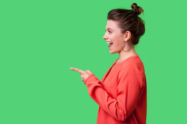 Photo of Hey you! Side view portrait of happy girl with bun hairstyle, big earrings and in red blouse. indoor studio shot isolated on green background
